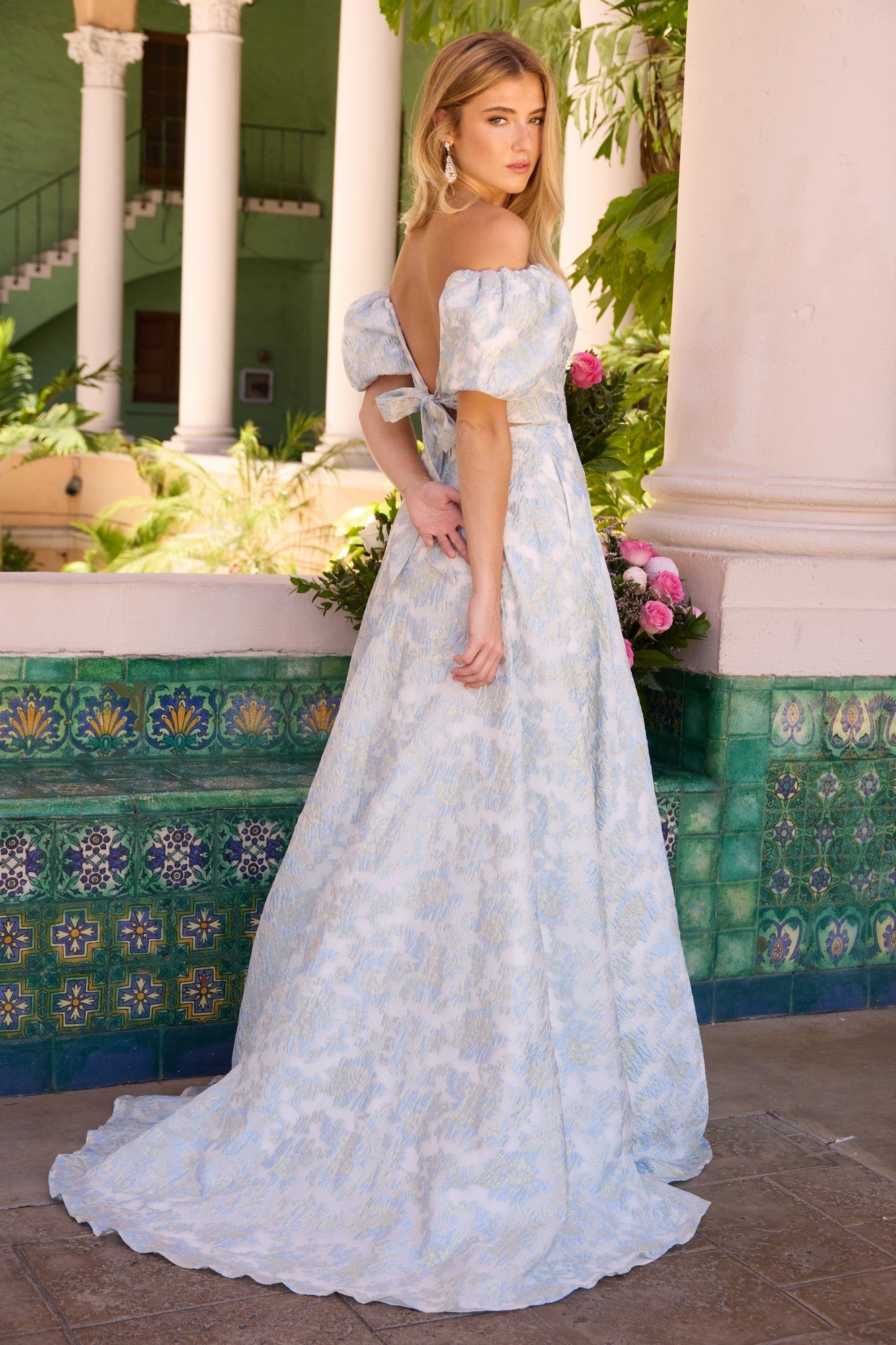 A woman wearing the Ava Presley 42267 long prom dress in light blue and white brocade with puff sleeves stands on a tiled patio near a column and greenery, glancing over her shoulder.